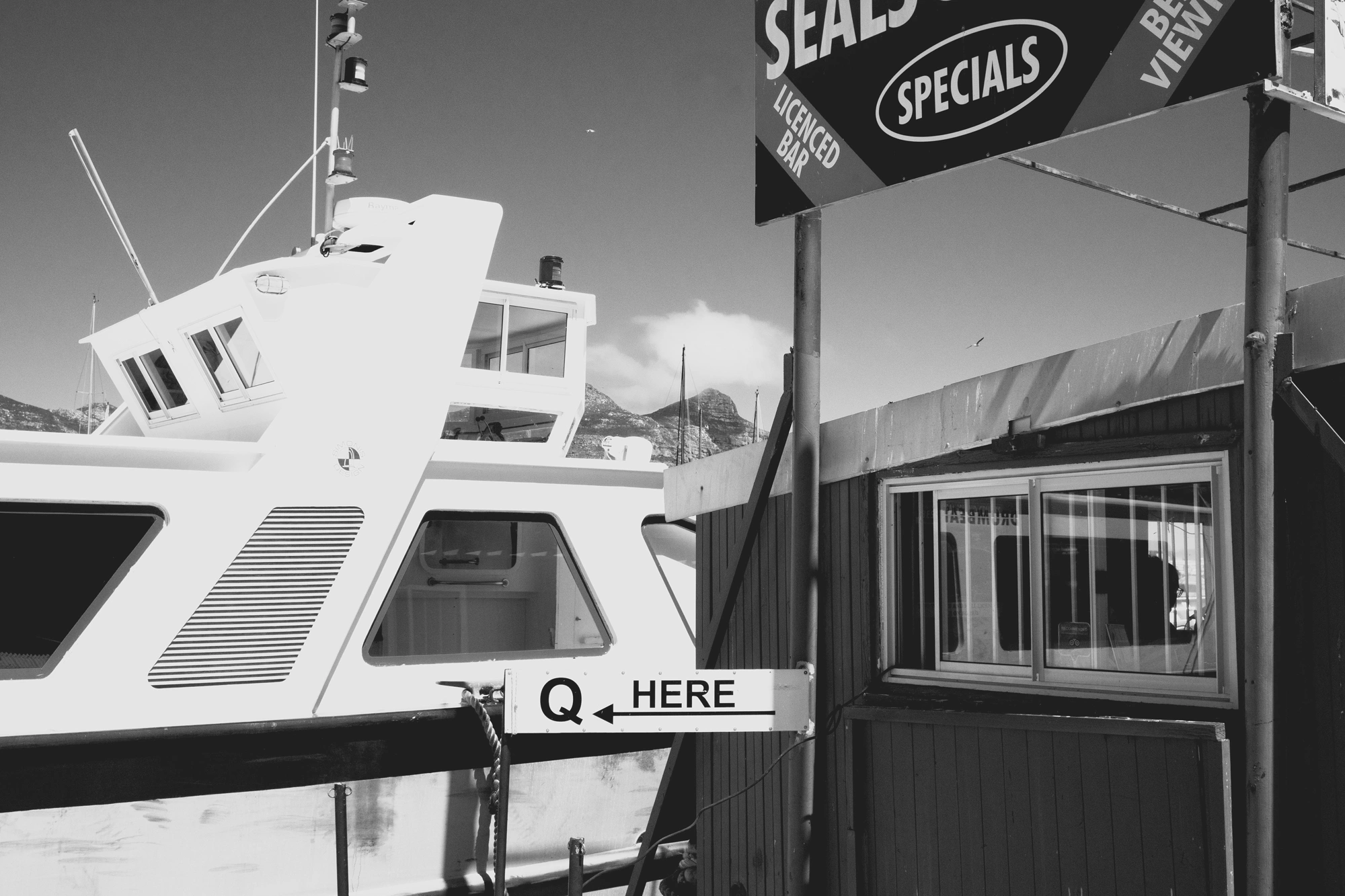 2022-02-13 - xt15-45 - bw - ZA - Cape Town - A ticket booth beside a boat with a sign reading 'Q Here'
