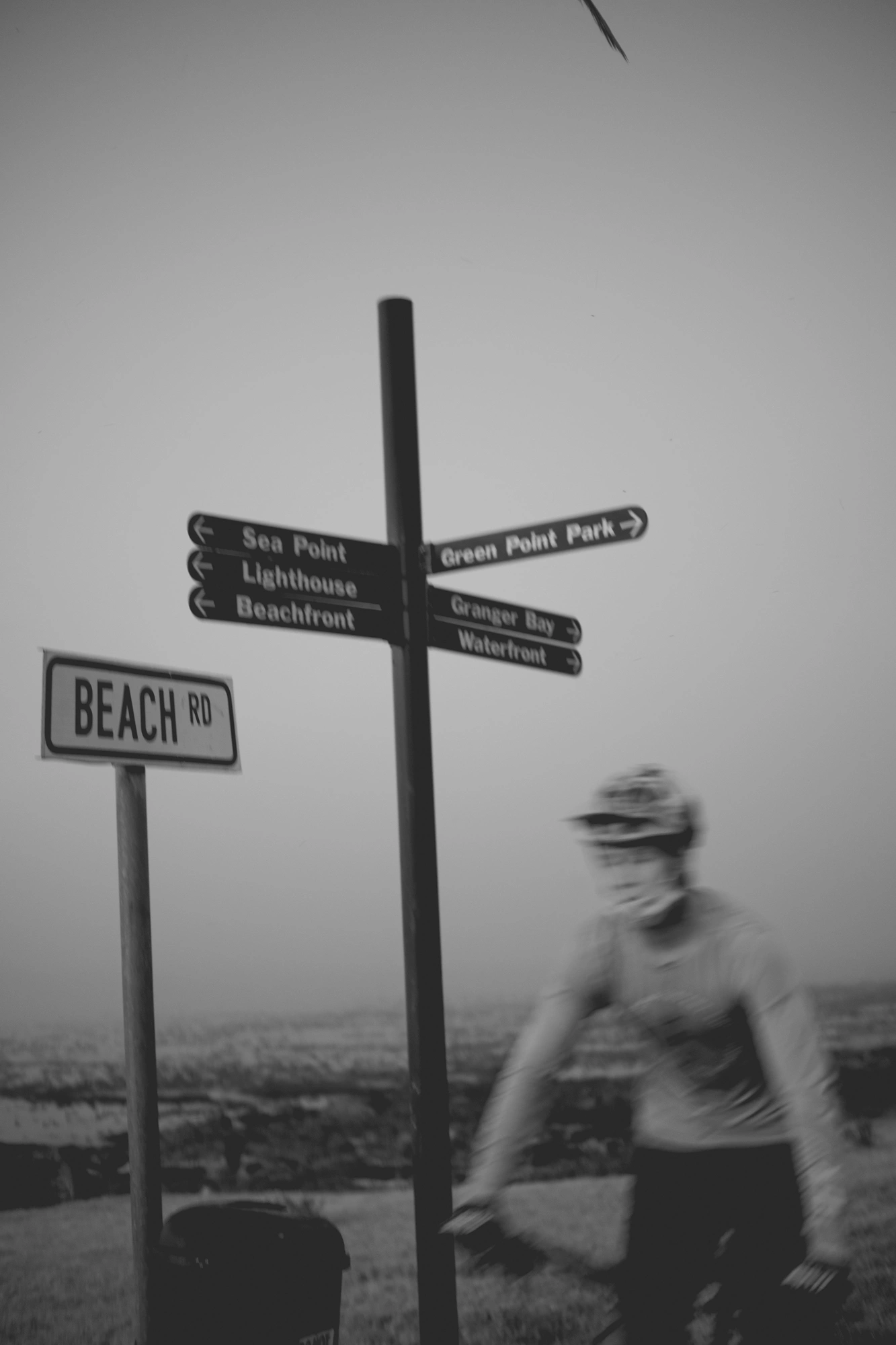 2022-02-15 - xt35 - bw - ZA - Cape Town - Man rides in front of sign that reads 'Beach Road'
