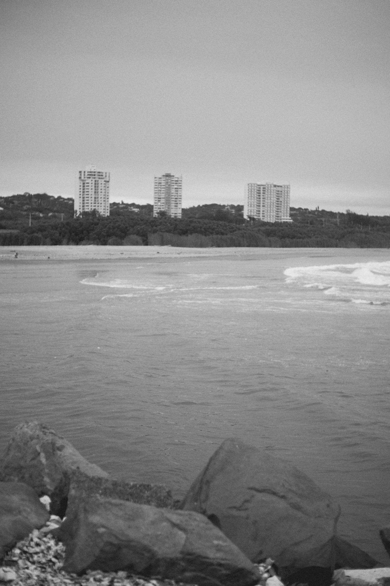 2022-09-21 - xt35 - bw - ZA - Durban - Rocks with buildings in background over the ocean
