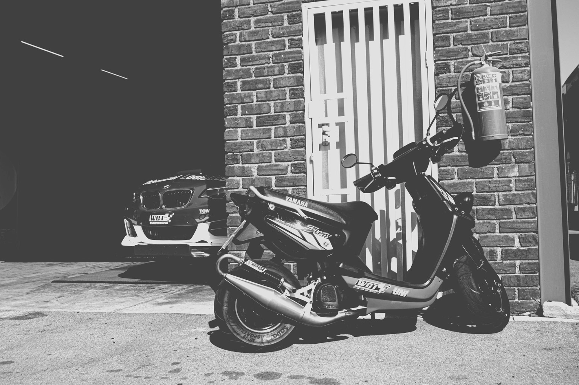 2019-07-27 - n18-55 - bw - ZA - Zwartkops Raceway - Scooter parked next to garage with race car 