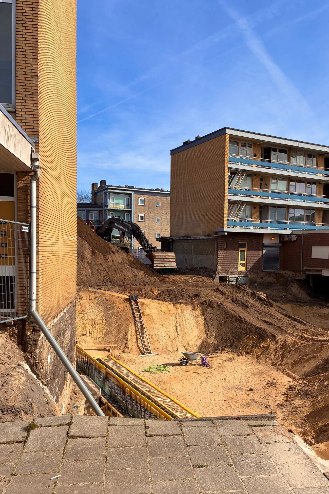 2026-03-20 - Big hole being dug by excavator between appartment buildings.J